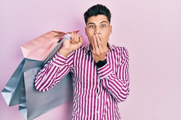 Canvas Print - Young hispanic man holding shopping bags covering mouth with hand, shocked and afraid for mistake. surprised expression