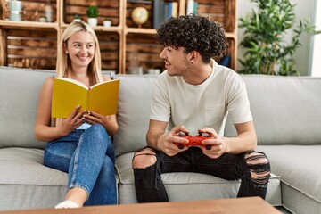 Sticker - Young couple smiling happy playing video game and reading book at home.