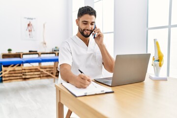 Sticker - Young arab man wearing physiotherapist uniform talking on the smartphone at clinic