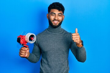 Wall Mural - Arab man with beard holding packing tape smiling happy and positive, thumb up doing excellent and approval sign