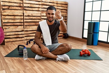 Poster - Young indian man sitting on training mat at the gym angry and mad raising fist frustrated and furious while shouting with anger. rage and aggressive concept.