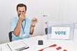 Middle age man with beard sitting by ballot holding i vote badge smelling something stinky and disgusting, intolerable smell, holding breath with fingers on nose. bad smell