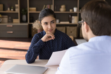 Canvas Print - Interested young professional female Indian human resources manager holding paper CV resume in hands, listening skilled male Caucasian job candidate at interview meeting, employment hiring concept.