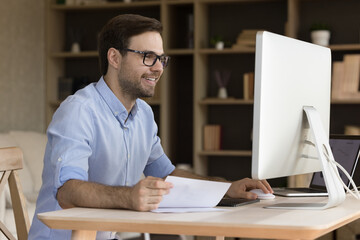 Happy young businessman in eyeglasses working on computer, analyzing paper document, feeling excited analyzing economic research report, satisfied with results or data statistics in modern home office