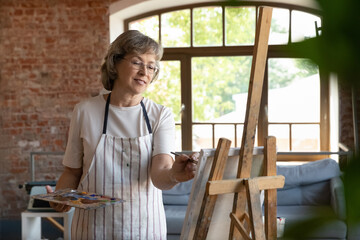 Happy senior retired lady drawing in paints on canvas. Elder talented professional artist working at easel in artistic studio, using palette, paintbrush, smiling, enjoying creativity, inspiration