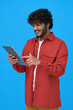 © insta_photos - Happy indian young man using digital tablet isolated on blue background. Smiling ethnic hipster guy holding pad playing game in app, buying online in ecommerce store, reading ebook. Vertical