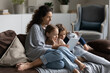 Loving young mom reading book interesting fairy tale story to little 5s adorable son and 6s daughter, family enjoy leisure time sit on cozy sofa at modern home. Develop kids, babysitting work concept