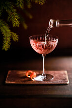 Elegant Coup Glass With Pink Drink And Orange Spiral. Rich Brown Tones For Surface And Background With Christmas Foliage In The Corner. Rose Pouring Into Glass From The Right.