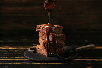 Pouring of chocolate topping onto tasty brownie pieces on black wooden background