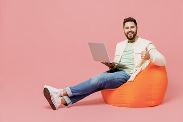 Wall Mural - Full body young smiling man in trendy jacket shirt sit in bag chair hold use work on laptop pc computer show thumb up isolated on plain pastel light pink background studio. People lifestyle concept.