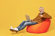 Full size body length elderly gray-haired bearded man 40s years old wears brown shirt sit in bag chair hold use point finger on laptop pc computer isolated on plain yellow background studio portrait.