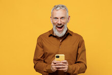 Shocked Surprised Happy Elderly Gray-haired Bearded Man 40s Years Old Wears Brown Shirt Look Camera Hold Use Mobile Cell Phone Keep Mouth Wide Open Isolated On Plain Yellow Background Studio Portrait.