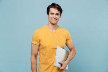 Young smiling confident programmer happy man 20s in yellow t-shirt hold closed laptop pc computer look camera isolated on plain pastel light blue background studio portrait. People lifestyle concept