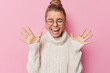 Leinwandbild Motiv Horizontal shot of happy woman with combed hair raises palms wears round spectacles and white sweater feels amused poses against pink background hears amazing good news screams from happiness