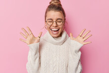 Horizontal Shot Of Happy Woman With Combed Hair Raises Palms Wears Round Spectacles And White Sweater Feels Amused Poses Against Pink Background Hears Amazing Good News Screams From Happiness