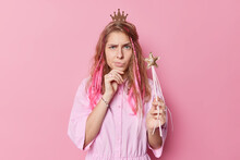Upset Discontent Beautiful Young Woman Has Sulking Expression Raises Eyebrows Wears Princess Crown And Dress Holds Magic Wand Ready To Make Your Dreams Come True Isolated Over Pink Background.