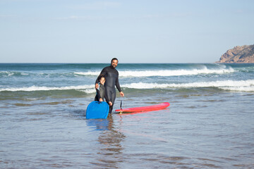 Pleased father and daughter doing sports on beach. Mid adult man and little dark-haired girl standing in water with surfboards, father hugging child. Family, leisure, active lifestyle concept