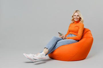 Full body elderly smiling happy blonde woman 50s in orange turtleneck sit in bag chair hold in hand use mobile cell phone isolated on plain grey background studio portrait. People lifestyle concept.