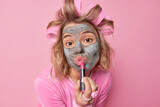 Fototapeta  - Portrait of lovely young European woman applies lipstick beauty nourishing mask and hair rollers prepares for date wants to look beautiful uses cosmetic products isolated over pink background.