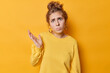 Confused doubtful young European woman with combed hair raises palm and looks puzzled reacts on something unexpected wears casual jumper poses against vivid yellow background. So what to do now