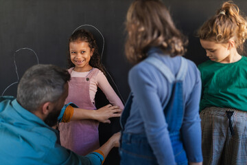 Wall Mural - Teacher drawing little girl student with chalks on blackboard wall indoors in playroom.