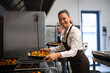 Happy female cook looking at camera and holding tray with baked pumpkin pieces in commercial kitchen