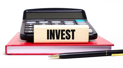 Fototapete - A burgundy notebook on a white background. On it is a black calculator, a pen and a wooden block with a INVEST text. Business concept