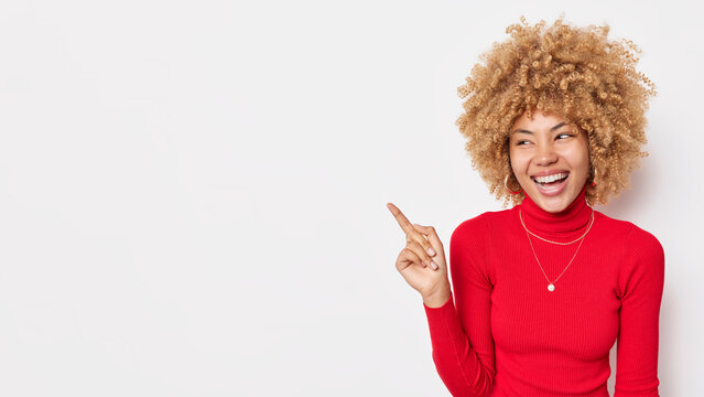 Positive happy woman with curly bushy hair points away at blank space shows advertisement or promotion wears red turtleneck feels glad isolated over white background blank copy space for text