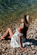 © Andrii - A beautiful young woman in a hat, glasses and a light dress is sitting with her back to the ocean against the background of huge rocks on a sunny day. Tourism and tourist trips.