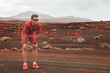 © Maridav - Tired sport athlete runner taking a break breathing during exercise workout on mountain trail volcanic landscape. Man wearing sunglasses, red compression clothes, smartwatch