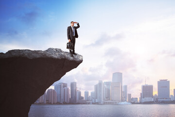 Canvas Print - Handsome young european man on cliff edge looking into the distance on bright city background with daylight and mock up place. Future, success, leadership and career growth concept.