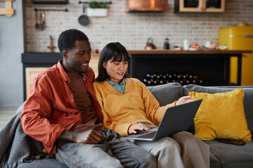 Portrait of young mixed-race couple using laptop while relaxing on couch in cozy home interior, copy space