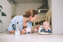 Loving smiling mother and baby eating breakfast and have fun in kitchen at home. Beautiful mother enjoys her life on maternity leave with her child.