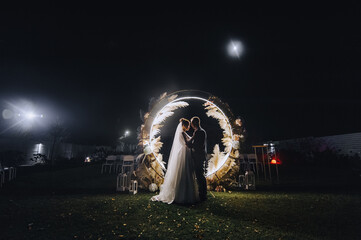 A stylish groom and a beautiful bride in a long dress are standing, hugging at night near a luminous cane arch decorated with lamps and garlands. Wedding photography, portrait.