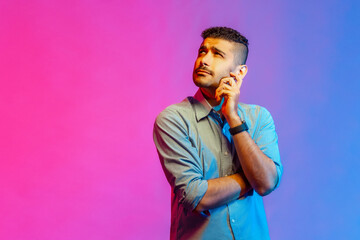 Wall Mural - Portrait of thoughtful handsome man in shirt holding his chin and pondering idea, confused not sure about solution. Indoor studio shot isolated on colorful neon light background.