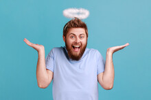 Portrait Of Bearded Angelic Man With Holy Nimbus Looking At Camera With Toothy Smile, Expressing Positive Emotions, Raised Arms, Looks Happy. Indoor Studio Shot Isolated On Blue Background.