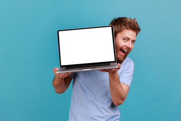 Wall Mural - portrait of bearded man holding laptop with blank white screen and looking at camera with excited fa