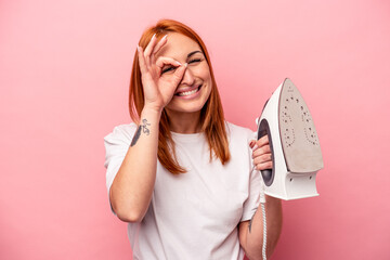 Young caucasian woman holding iron isolated on pink background excited keeping ok gesture on eye.