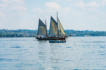 Wall Mural - Old wooden sailing ship and a motorboat in motion on the Lake Garda in front of the small town of Garda, tourist resort in Verona province, Veneto, Italy, southern Europe.