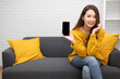 © comzeal - Young elegant beautiful Asian woman smiling and showing screen on mobile phone and sitting on sofa with laptop in the living room at home