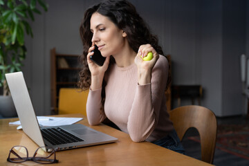 Wall Mural - Multitasking young businesswoman gripping rubber expander in hand while talking on mobile phone sitting at laptop computer at workplace. Female making exercises for tunnel syndrome treatment in office