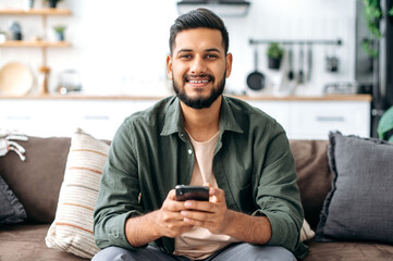 Happy handsome indian or arabian guy in casual wear, sits at home on sofa, using his smartphone, texting online, browsing social networks, messaging with friends, looks at camera, smiles friendly