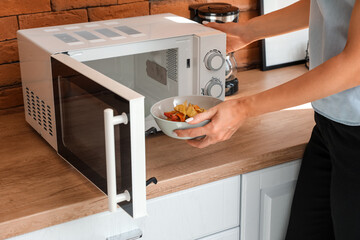 Woman putting bowl with delicious food into modern microwave oven on wooden counter near brick wall