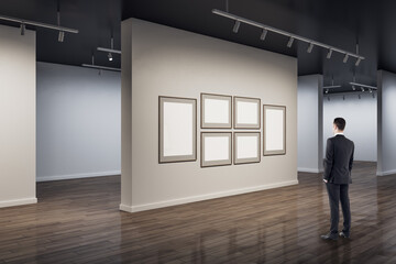 Poster - Businessman in modern concrete exhibition hall interior looking at blank white mock up posters on wall and wooden flooring with reflections. Art and museum concept.