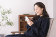 © lielos - ソファーに座りながらスマホを見ているアジア人女性、自宅リビング