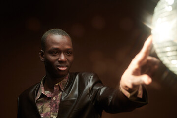 Wall Mural - Happy young African man touching discoball at disco in the night club while standing in front of camera over dark background