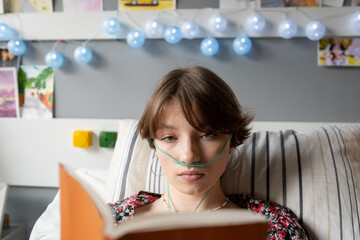 close-up of sick teenage girl with oxygen tube in her nose reading a book while lying on bed at hosp