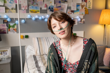 portrait of teenage girl breathing with special oxygen tube looking at camera while sitting on bed a