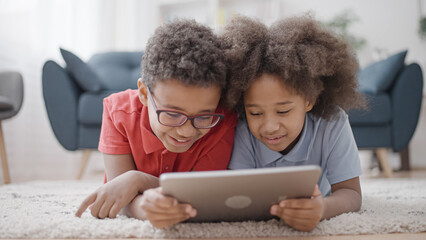 Wall Mural - Cheerful black siblings lying on floor with tablet, having fun together, smiling