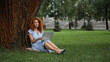 © LIGHTFIELD STUDIOS - full length of curly redhead woman in dress listening music while using laptop in park.